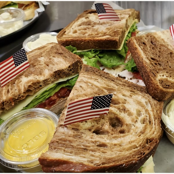 Four deli sandwiches on toasted bread with lettuce, cheese, and meat, each with an American flag pick.