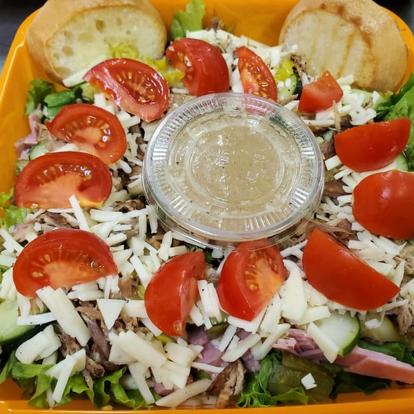 A deli salad with lettuce, tomatoes, shredded cheese, meat, and a side of dressing.