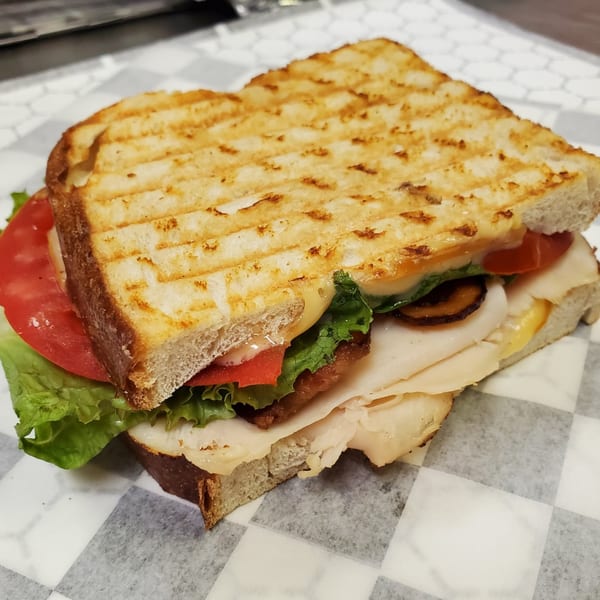 Grilled deli sandwich with turkey, cheese, lettuce, tomato, and bacon on checkered paper.