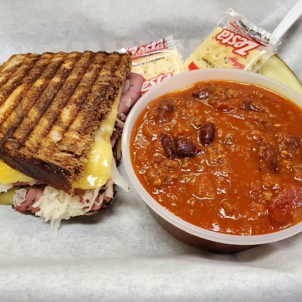 Grilled Reuben sandwich with melted cheese, sauerkraut, and pastrami, served with a side of chili and crackers.