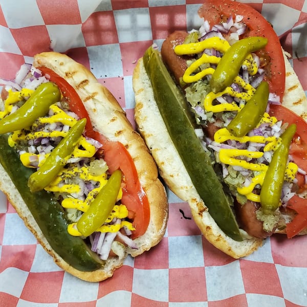 Two deli hot dogs with tomatoes, onions, relish, mustard, and peppers on checkered paper.