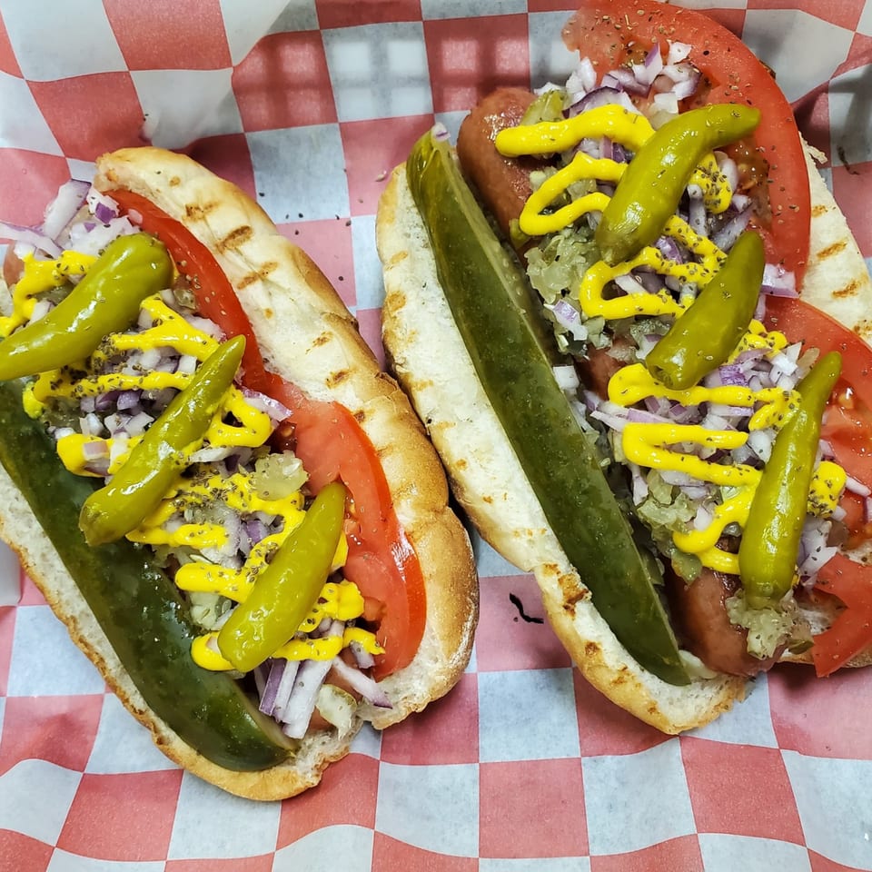 Two deli hot dogs with tomatoes, onions, relish, mustard, and peppers on checkered paper.