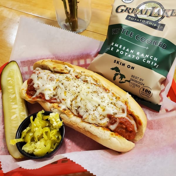 Meatball sub with melted cheese, a pickle spear, chopped peppers, and a bag of chips.