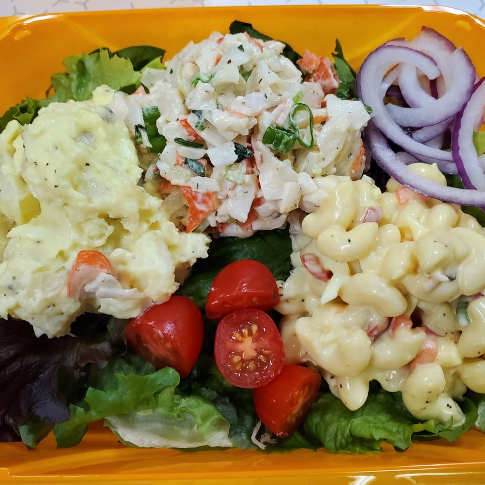 Deli salad platter with potato salad, crab salad, macaroni salad, cherry tomatoes, and red onion.